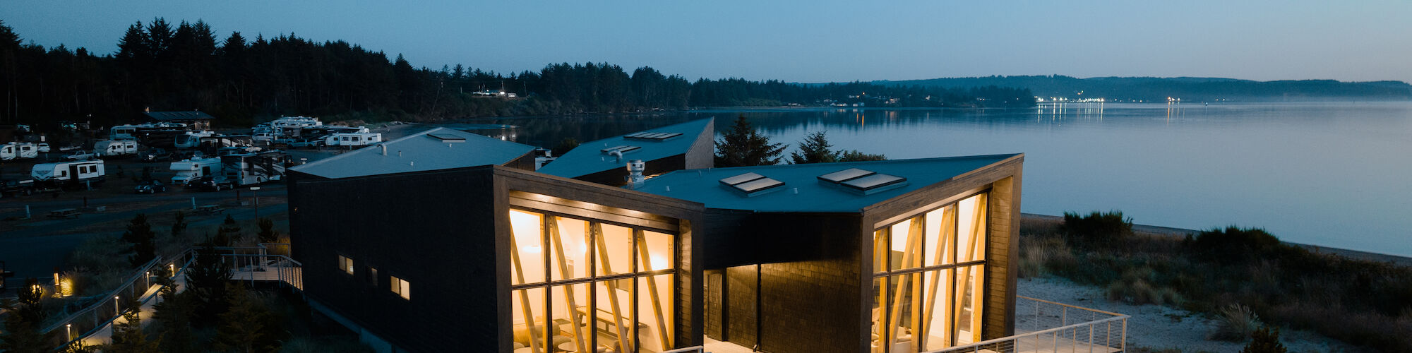 A modern resort clubhouse with illuminated windows is situated by the waterfront, surrounded by greenery, during twilight with a clear evening sky.