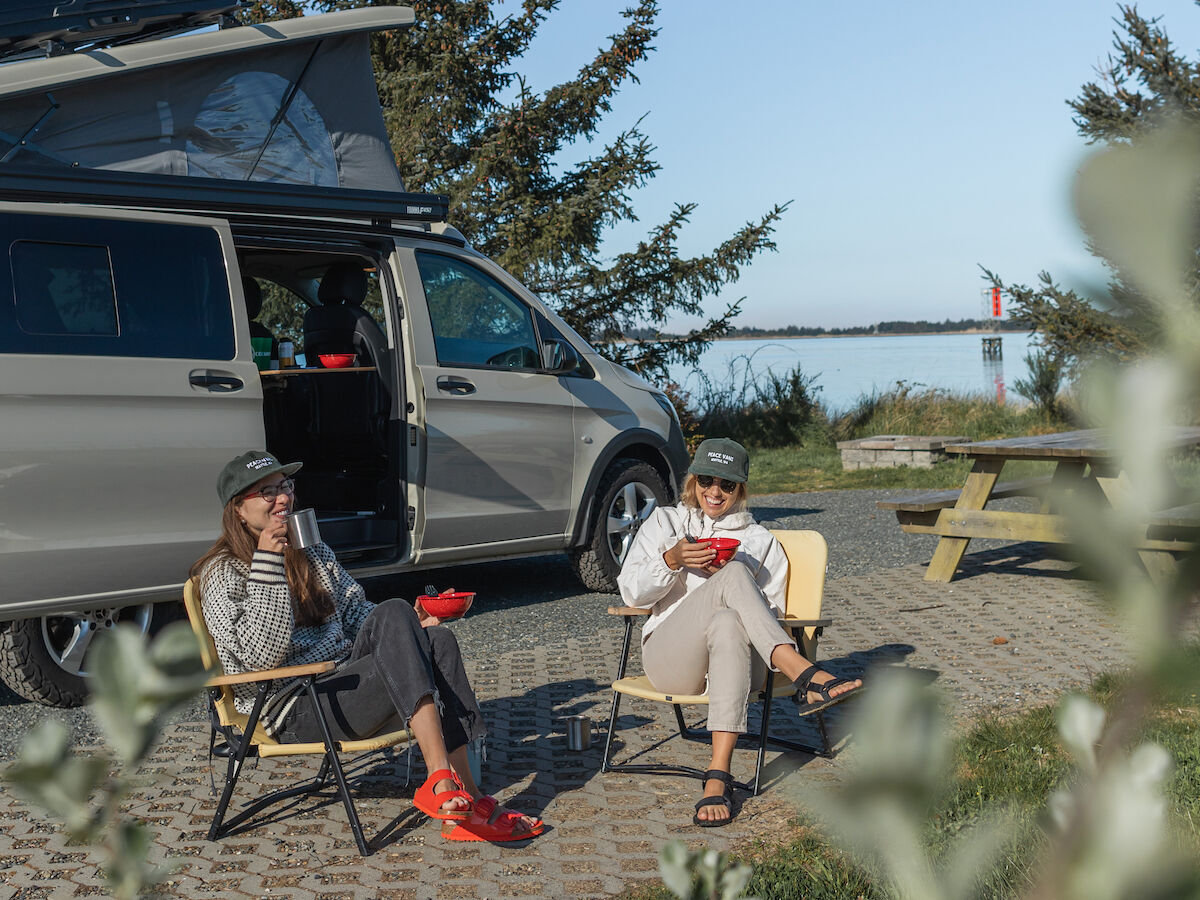 Two people relax in chairs near a parked van with an open roof box, surrounded by trees and a clear blue sky in the background.
