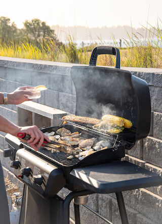 A person is grilling chicken and corn on a barbecue grill outdoors, with smoke rising and a scenic view in the background.