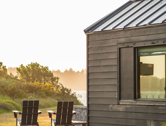 A modern house with a metal roof, wooden exterior, and window. Two wooden chairs on a deck, surrounded by grass and a scenic, sunlit landscape.