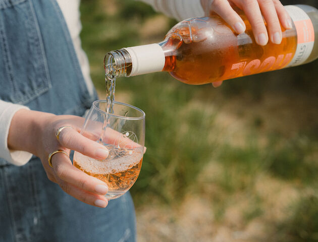 Someone is pouring rosé wine from a bottle into a clear glass outdoors, wearing denim overalls.