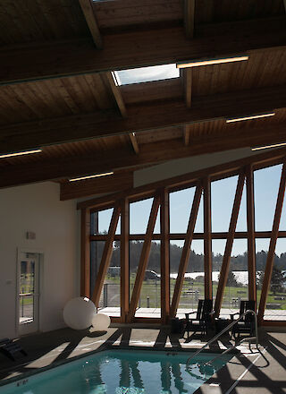 A modern indoor space with a pool, wooden beams, large windows, and poolside furniture, letting in natural light.