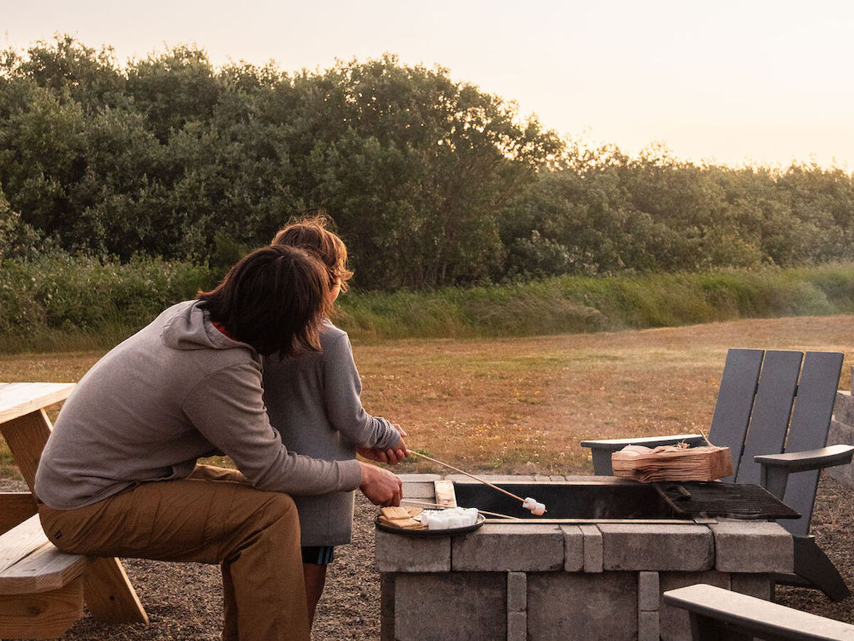 Two people sit near a fire pit outdoors, surrounded by chairs, under a clear sky at sunset.