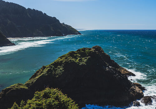 A scenic coastal view with blue waters, rugged cliffs, green vegetation, and a clear blue sky.