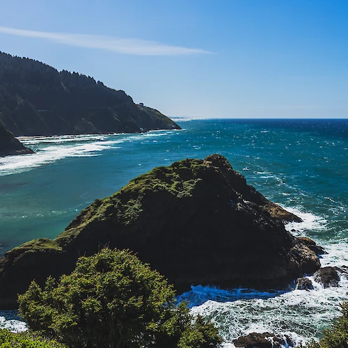 A scenic coastal view with blue waters, rugged cliffs, green vegetation, and a clear blue sky.