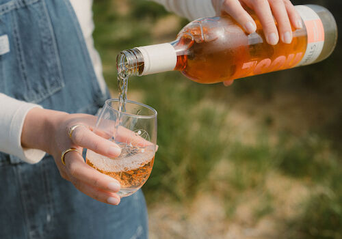 A person pours rose wine from a bottle into a glass outdoors, with a blurred natural background.