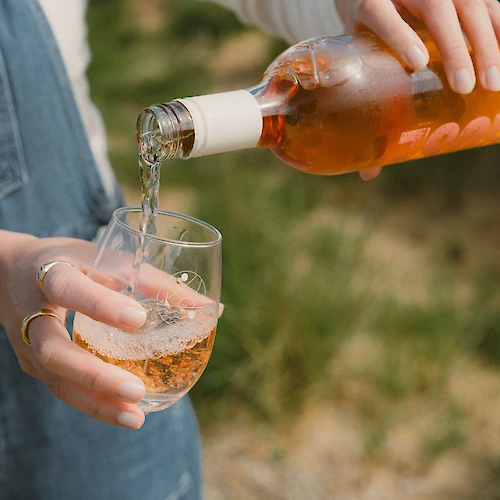 A person pours rose wine from a bottle into a glass outdoors, with a blurred natural background.