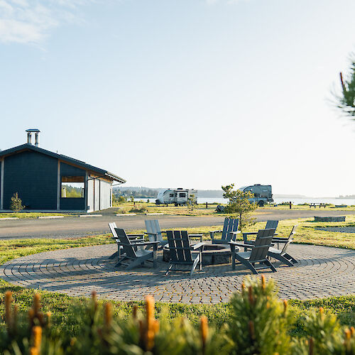 A patio with wooden chairs arranged in a circle, a small building, and scenery with trees and open sky in the background.