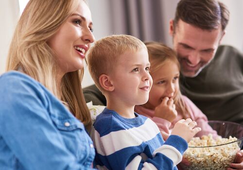 A family, smiling and relaxed, sits on a sofa together, watching something with a bowl of popcorn in their laps.