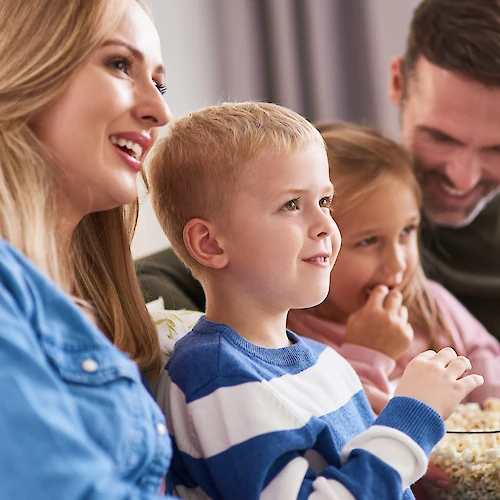 A family, smiling and relaxed, sits on a sofa together, watching something with a bowl of popcorn in their laps.