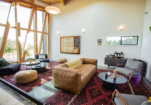 A modern living room features a large sofa, round tables, armchairs, and geometric windows on a patterned rug.