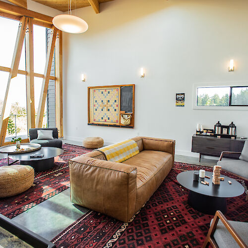 A modern living room features a large sofa, round tables, armchairs, and geometric windows on a patterned rug.