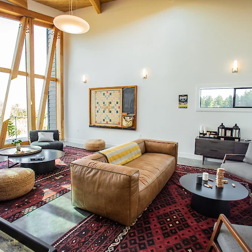 A modern living room features a large sofa, round tables, armchairs, and geometric windows on a patterned rug.