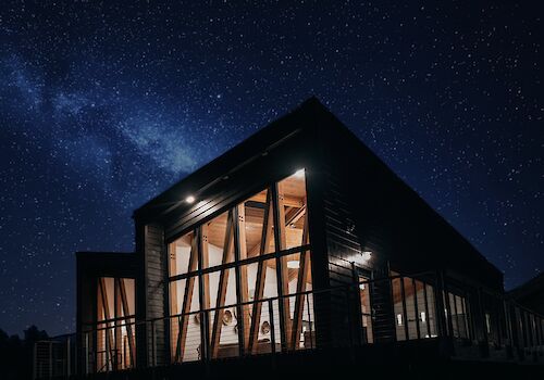 A modern, illuminated house with large windows under a starry night sky and Milky Way, creating a serene atmosphere.