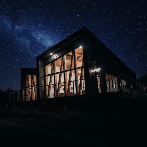 A modern, illuminated house with large windows under a starry night sky and Milky Way, creating a serene atmosphere.