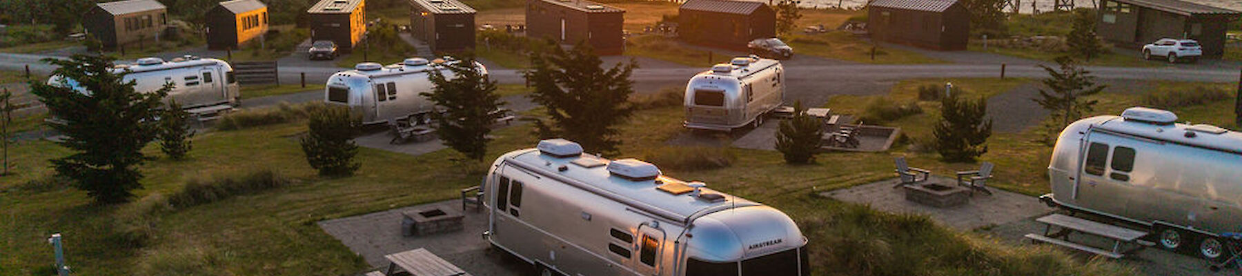 A serene RV park at sunset with multiple silver trailers, picnic tables, and trees, near a water body.