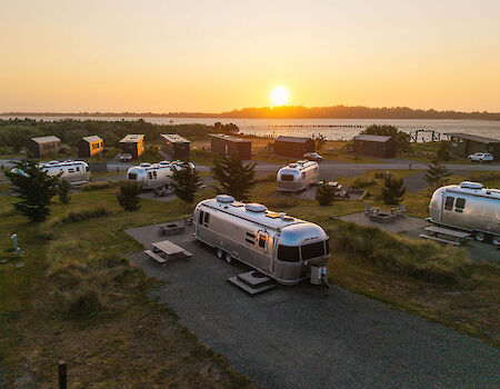 A serene RV park at sunset with multiple silver trailers, picnic tables, and trees, near a water body.