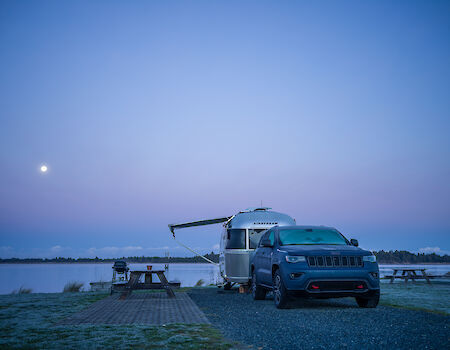 A car parked beside a camper near a lake, with a picnic table, under a twilight sky with the moon visible.