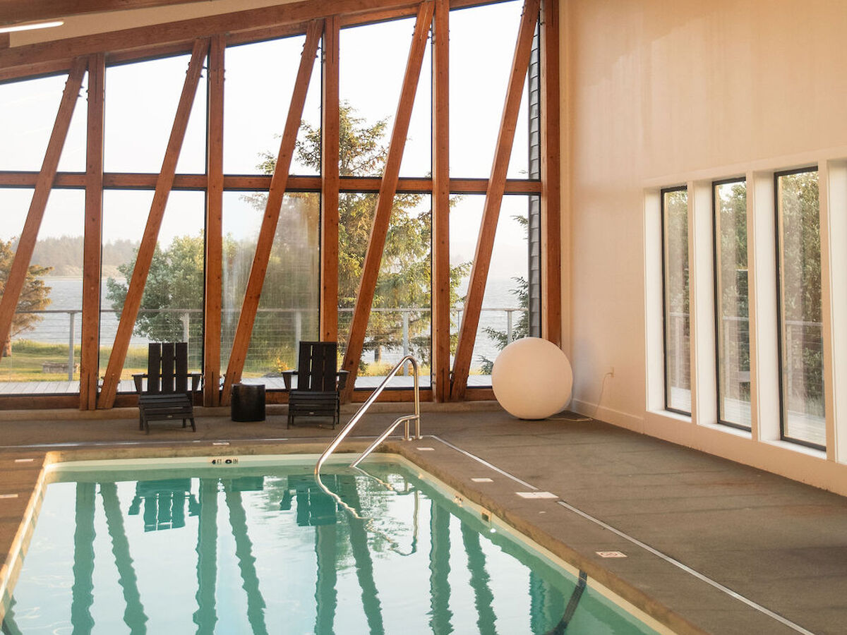 The image shows an indoor swimming pool with wooden beams, large windows, and a view of trees outside.