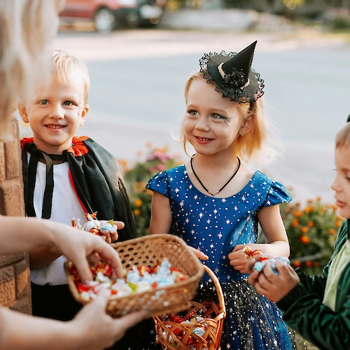 Three children in costumes enjoying trick-or-treating, receiving candy from an adult holding a basket full of treats.