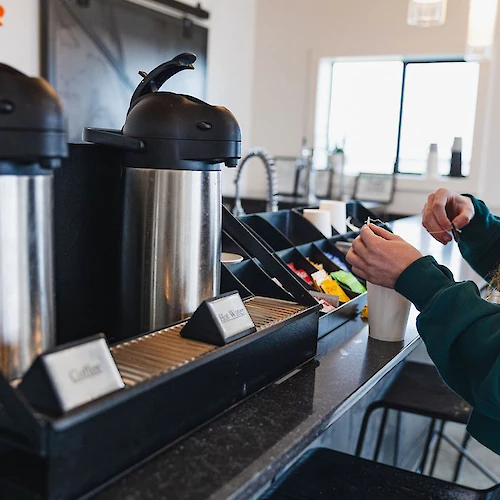 A person preparing a drink at a coffee station with thermoses, sugar packets, and cups in a cafe setting.