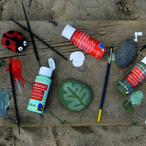 Painted stones resembling a ladybug and leaf, surrounded by paint bottles and brushes on a wooden surface.