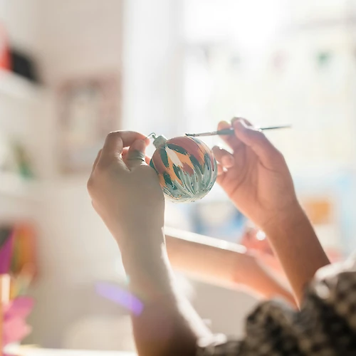 A person is painting a decorative object with a brush, sitting in a bright room with shelves and colorful paper in the background.