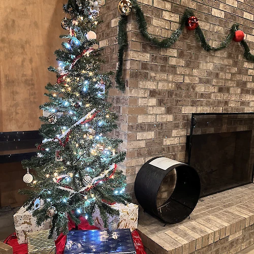 A decorated Christmas tree with lights, ornaments, and presents is next to a brick fireplace adorned with garlands and baubles.