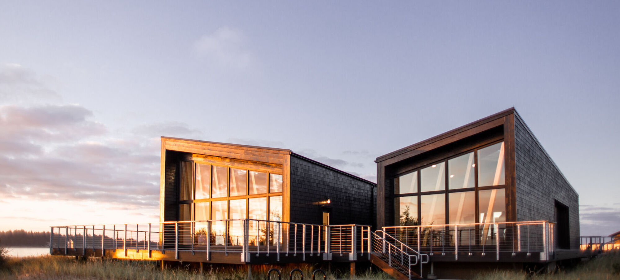 A modern building with large glass windows, surrounded by wooden decking, set against a twilight sky.