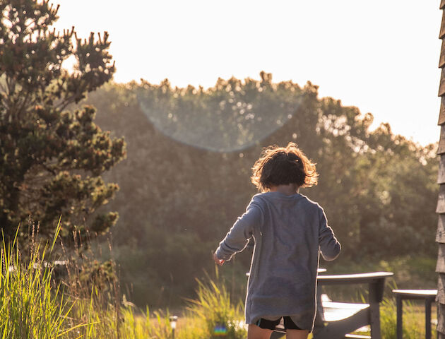 A person walking barefoot on a sunlit path by a house, surrounded by grass and trees, with a bright sky in the background.