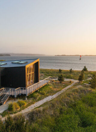A modern house with large windows is situated near a calm body of water, surrounded by greenery under a clear sky at sunset.