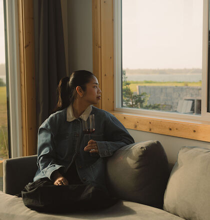 A person in a denim jacket is sitting on a couch, looking out a large window with a view of the outdoors and holding a phone.