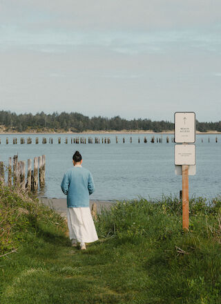 A person walking on a grassy path toward a body of water, with a sign in the foreground and trees in the distance.