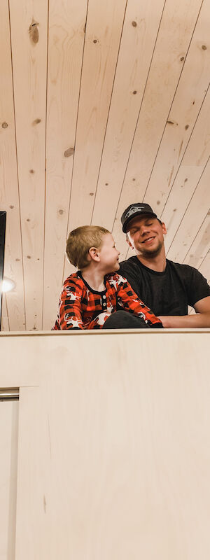 A person and a child in pajamas are sitting on a loft railing in a wood-paneled room, smiling at each other.