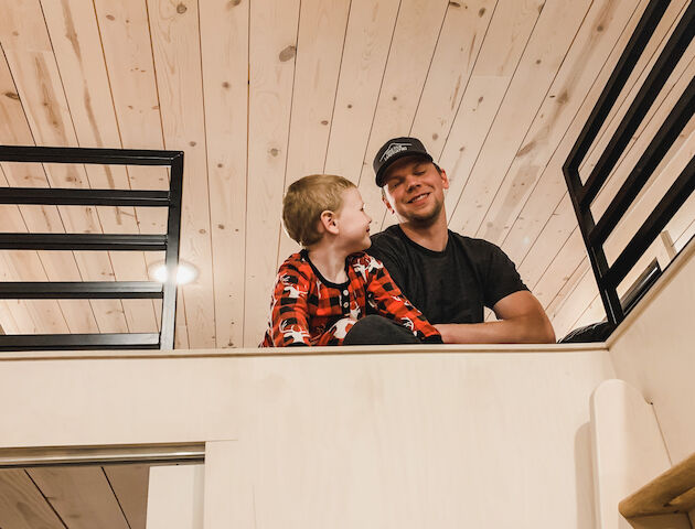 A person and a child in pajamas are sitting on a loft railing in a wood-paneled room, smiling at each other.