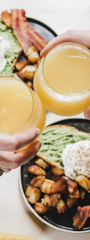 Two people clinking glasses of orange juice over a table with avocado toast, eggs, bacon, potatoes, bananas, and a mug.
