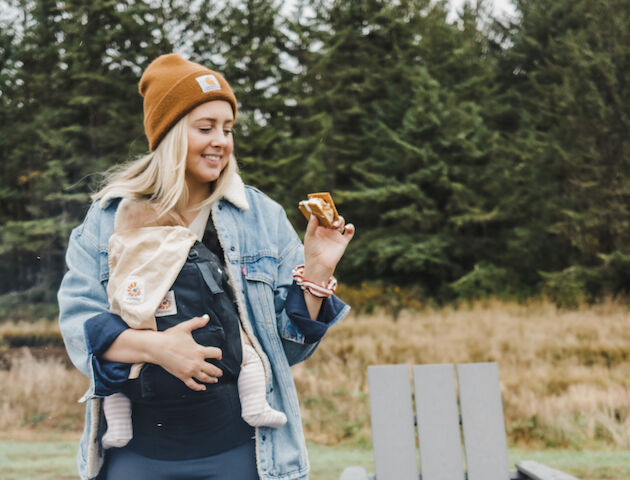 A person outdoors holding food, with a baby in a carrier, wearing a beanie and denim jacket, surrounded by trees.