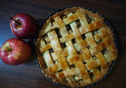 A lattice-topped apple pie sits next to two red apples on a dark wooden table.