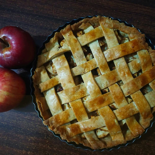 A lattice-topped apple pie sits next to two red apples on a dark wooden table.