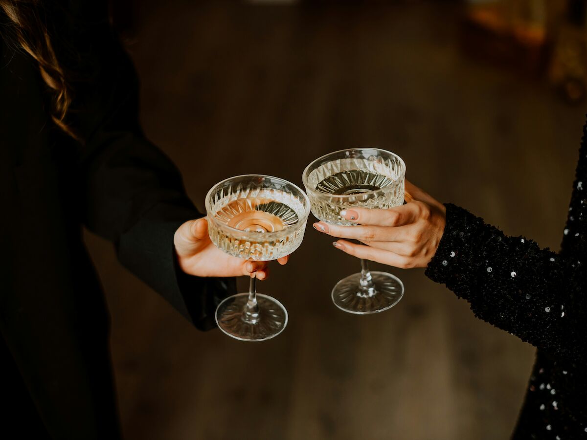 Two people clink crystal cocktail glasses in a toast, hands visible against a dark background, festive and elegant scene.
