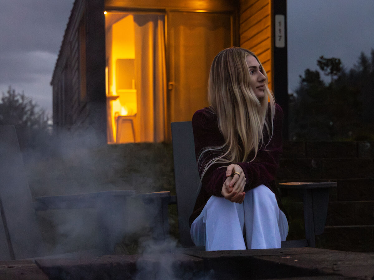 A woman sits by a small wooden cabin glowing warm inside, steam rising from a nearby fire, twilight sky above.