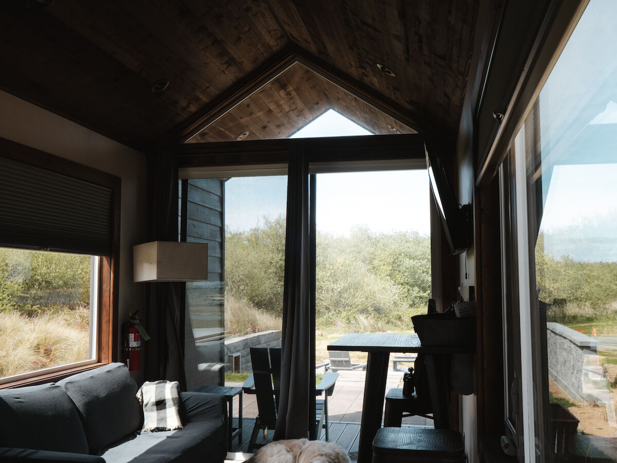 Cozy cabin interior with a sofa, lamp, and large windows; a dog lying on the floor by the glass wall, and a view of outdoor desert scenery.