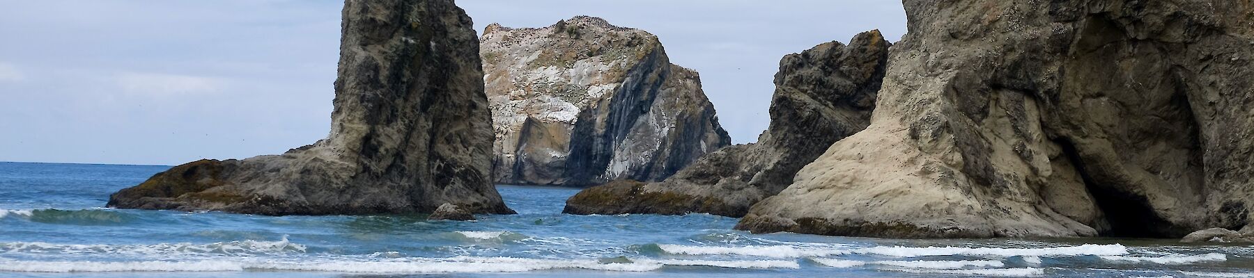 A rocky shoreline with large sea stacks rising from the ocean, waves gently lapping the sandy beach under a blue, partly cloudy sky.