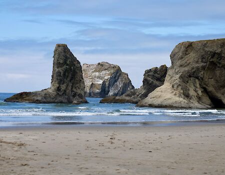 A rocky shoreline with large sea stacks rising from the ocean, waves gently lapping the sandy beach under a blue, partly cloudy sky.