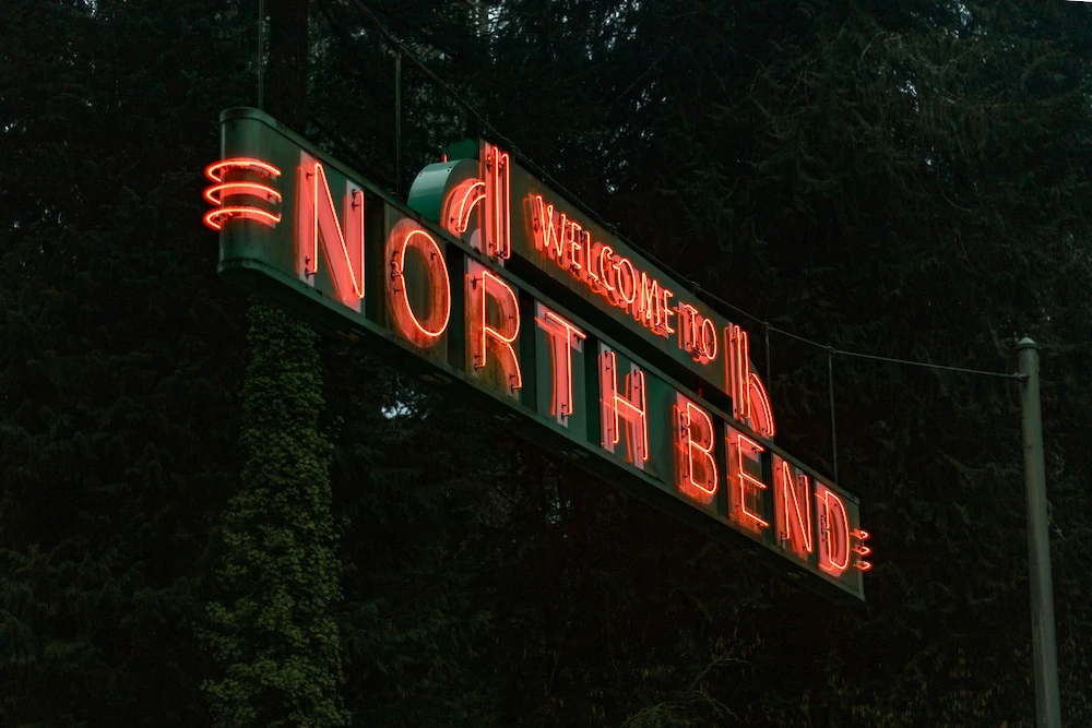 A neon sign reads &ldquo;WELCOME TO THE NORTH BEND&rdquo; in red/orange lights, mounted on a dark backdrop with trees behind it.