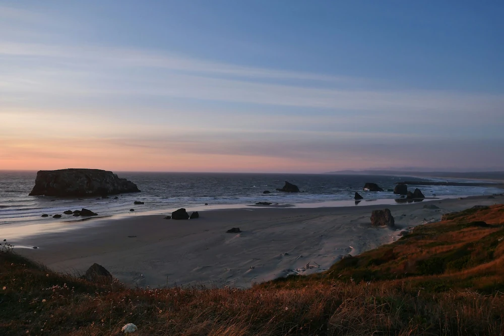 A serene coastal sunset over a rocky beach with calm waves, warm tones on the horizon, and grassy cliffs framing the shoreline.
