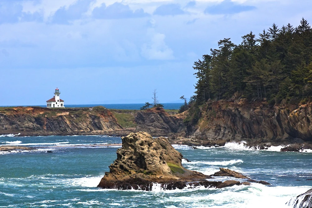 A rocky coastline with crashing waves, a lighthouse on a distant cliff, and pine-covered cliffs along the shore.