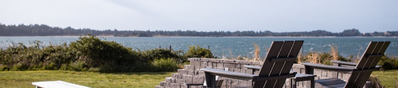 Sunlit patio by a calm lake with several black reclining lounge chairs and benches on a stone terrace, peaceful outdoors.