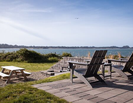 Sunlit patio by a calm lake with several black reclining lounge chairs and benches on a stone terrace, peaceful outdoors.