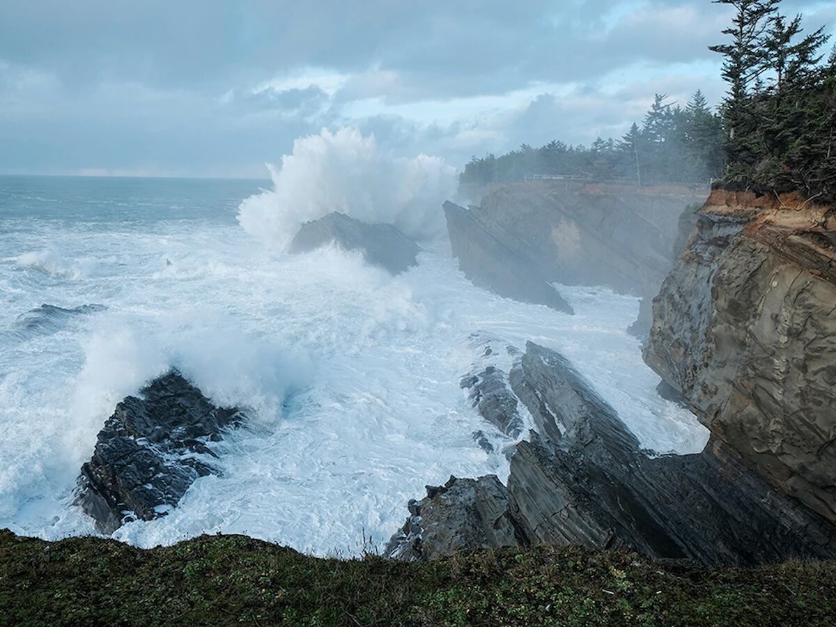 Rough ocean waves crash against rocky cliffs beside evergreen trees under a cloudy sky.